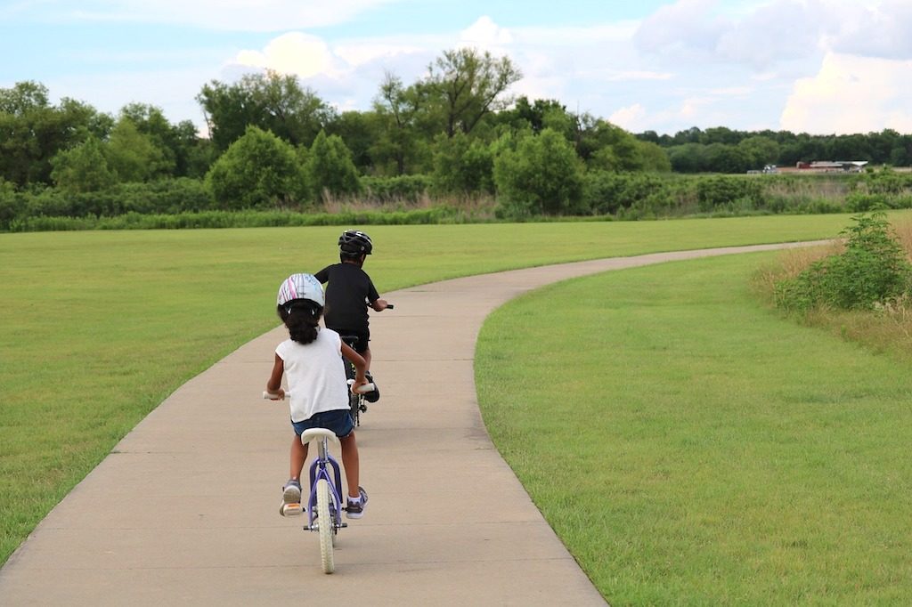 Two children riding bikes on a sidewalk in a park with trees in the background